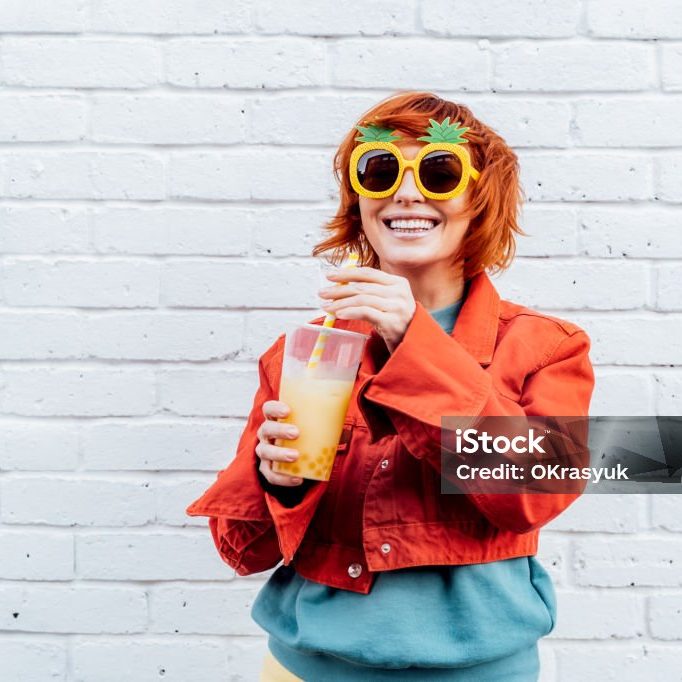 Hipster fashion young woman in bright clothes and pineapple glasses drinking fruity sugar flavored tapioca pearl bubble tea with straw on the white brick wall background. Selective focus. Copy space