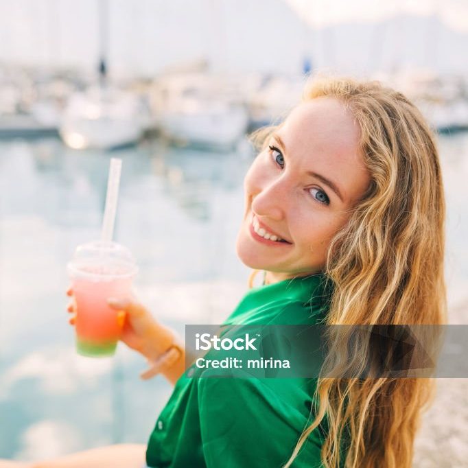 Blonde woman with long, wavy hair smiles while holding vibrant bubble tea drink, sitting by the water near boats at sunset.